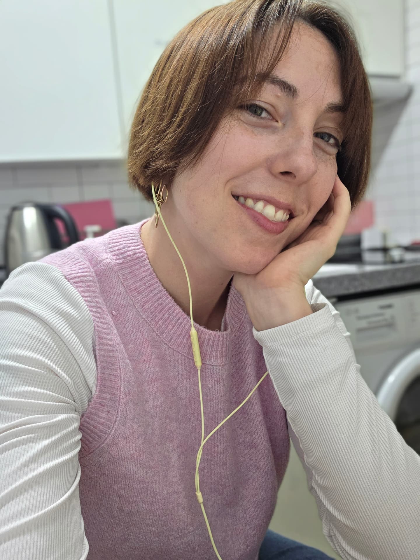 Abi resting her chin on her hand in a kitchen, wearing a pink knit vest and yellow earbuds